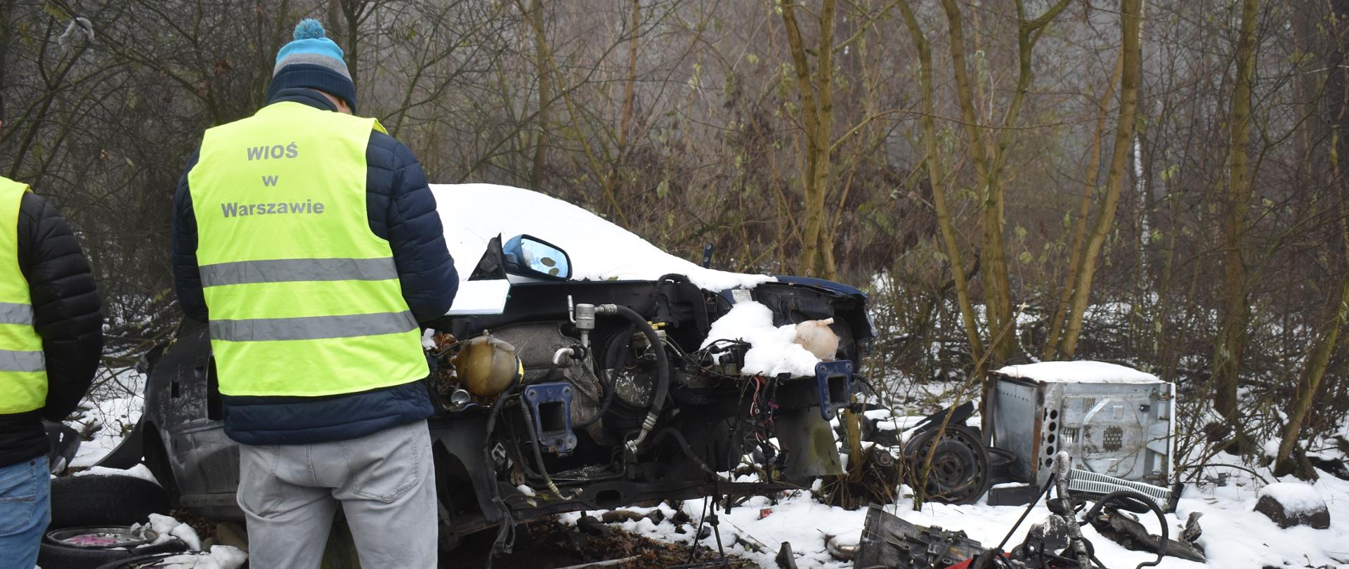 Inspektor WIOŚ w Warszawie w żółtej kamizelce odblaskowej prowadzi oględziny wraku rozmontowanego pojazdu na zaśnieżonym, zalesionym terenie; wokół leżą części samochodowe i opony.