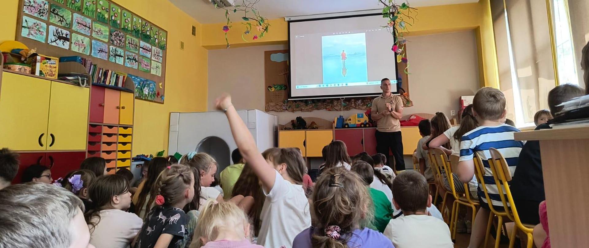 Strażak w sali lekcjyjnej opowiada dzieciom, włączona prezentacja na projektorze multimedialnym. Jedna z dziewczynek podnosi rękę. Sala bardzo kolorowa i upiększona.