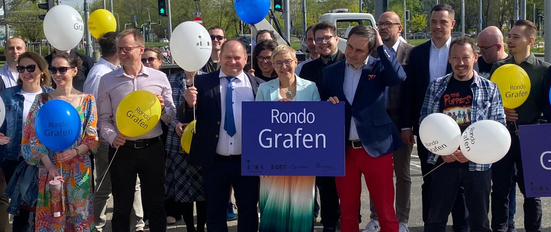 Na dworze, w słońcu, na chodniku wyłożonym szarą kostką stoi grupa ludzi, pośrodku wiceminister Mrówczyńska trzyma niebieską tablicę z napisem Rondo Grafen, w tle widać sygnalizację świetlną i zielone drzewa.