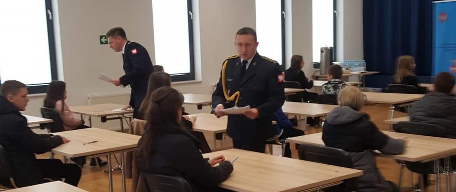 Na zdjęciu widać młodzież szkolną siedzącą w pomieszczeniu w pojedynczych ławkach ustawionych w rzędy. Pomiędzy ławkami widać dwóch strażaków państwowej straży pożarnej ubranych w mundury galowe rozdających młodzieży kartki z testami. 