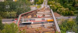 Zdjęcie ukazuje plac budowy wiaduktu nad czynną drogą. Na pierwszym planie znajdują się maszyny budowlane oraz materiały, takie jak rury i palety. Centralnie widoczna jest konstrukcja wiaduktu wsparta na betonowych filarach i stalowych belkach. Po obu stronach drogi ustawiono barierki ochronne i znaki ostrzegawcze. W tle widać jadące samochody, zabudowę mieszkalną oraz tereny zielone.