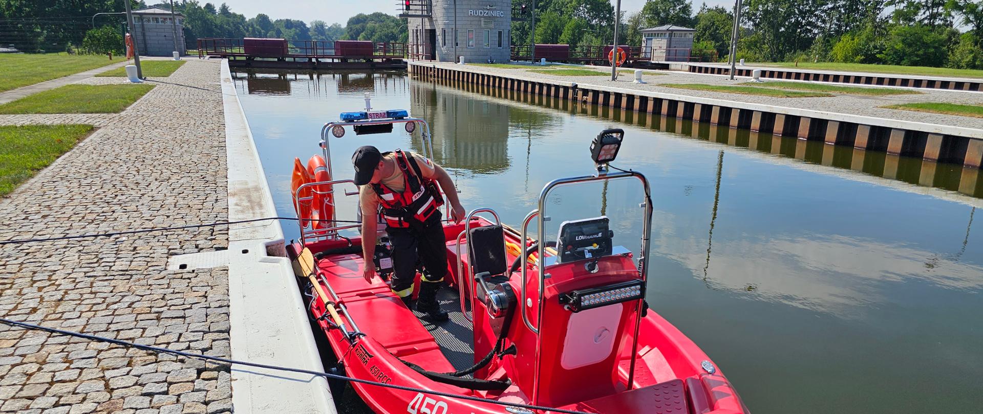 na zdjęciu widoczna czerwona, motorowa łódź ratownicza straży pożarnej. Łódź jest zacumowana na kanale gliwickim - przy śluzie Rudzinie. Na łodzi znajduje się strażak ochotniczej straży pożarnej. W tle budynki śluzy Rudziniec.