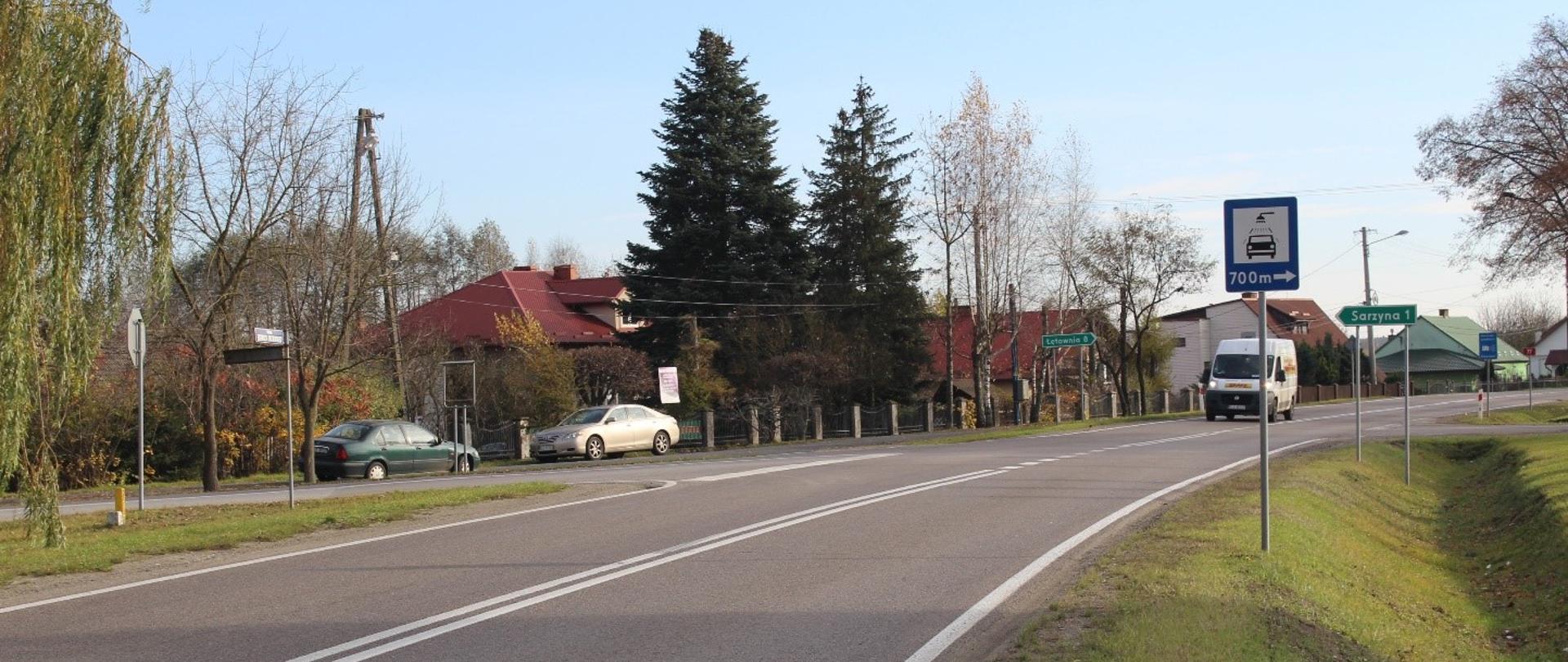 Widok na drogę krajową numer 77 w miejscowości Nowa Sarzyna, lekki łuk drogi w prawo. Jezdnia z dwoma pasami ruchu, po jednym pasie w każdym kierunku jazdy. Pasy ruchu rozdzielone podwójną linią ciągłą. Po prawej stronie widać znaki usytuowane na poboczu. Na jezdni jedzie bus, po lewej stronie zaparkowane dwa auta.