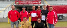 Grupa dziewięciu mężczyzn pozuje na bieżni stadionu przed trybunami. Większość ubrana jest w czerwone bluzy sportowe, jedna osoba ma beżowy mundur strażacki i trzyma puchar, a dwie osoby trzymają dyplomy. W tle widoczny jest czerwony baner informujący o mistrzostwach w sporcie pożarniczym Państwowej Straży Pożarnej województwa mazowieckiego. Zdjęcie przedstawia ciechanowskich strażaków podczas ceremonii pamiątkowej na stadionie.