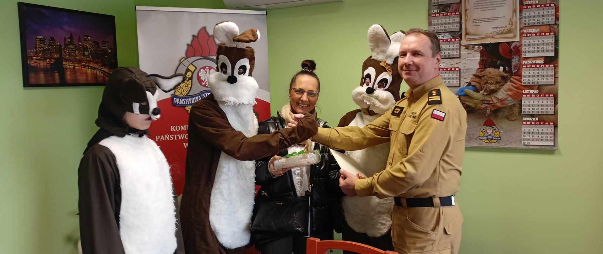 W gabinecie Komendanta Powiatowego przedstawiciele DPS w Zdunach składają wielkanocne życzenia. Przedstawiciele DPS w strojach zajączka wielkanocnego składają na ręce przedstawiciela strażaków kartkę z życzeniami oraz słodki upominek. Komendant Powiatowy ściska dłoń zajączka w podziękowaniu za świąteczną wizytę