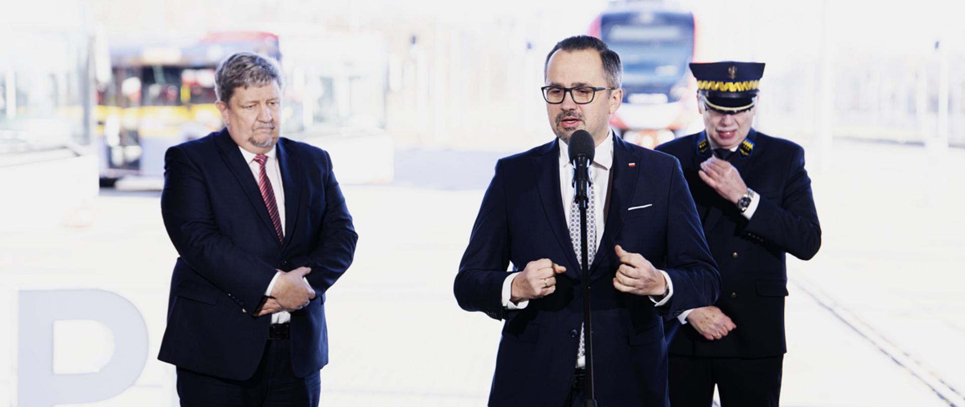 Trzy osoby stoją obok siebie na scenie. W środku do mikrofonu mówi wiceminister Marcin Horała.
