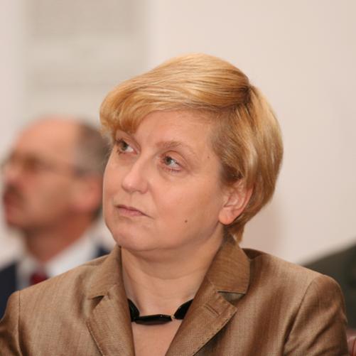 Anna Fotyga kolorowy portret kobiety, krótkie blond włosy, złota marynarka, czarne korale, w tle zamglone postaci mężczyzn.