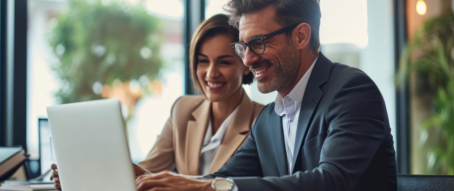 Na zdjęciu widać uśmiechniętych ludzi: pana i panią, którzy patrzą w laptop. 