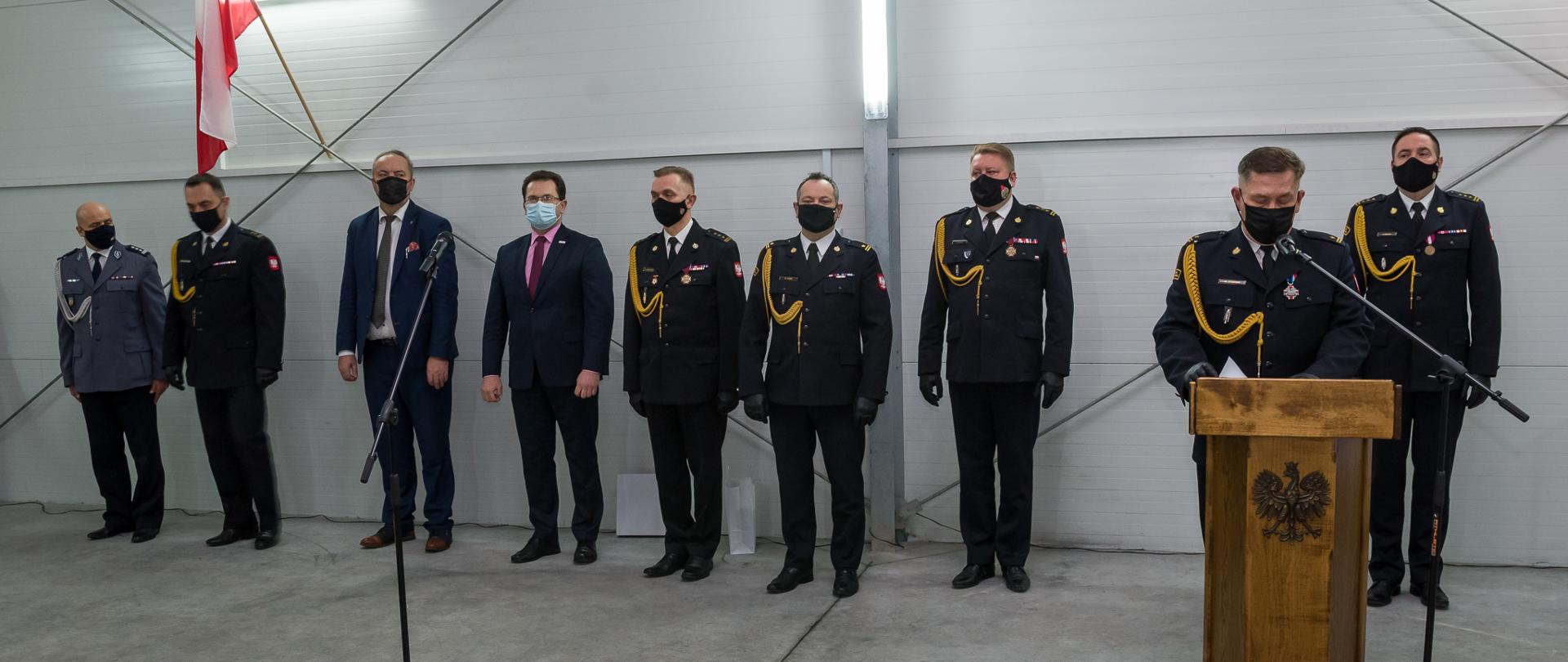 na zdjęciu sześciu strażaków i jeden policjant w mundurach galowych oraz dwóch mężczyzn w garniturach stojących w hali w rzędzie pod ścianą, jeden ze strażaków stoi przy mównicy, na ścianie hali flaga polski