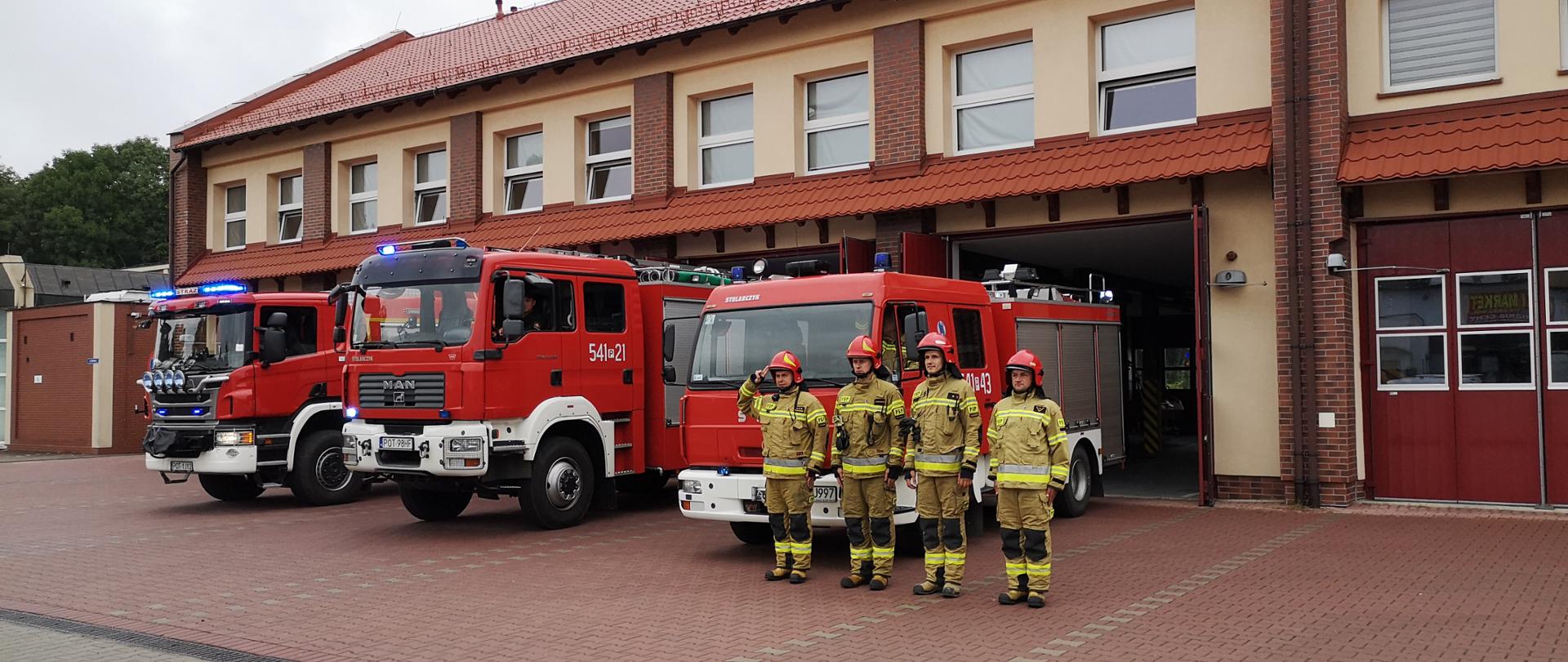 Na zdjęciu strażacy w trakcie oddawania hołdu Powstańcom Warszawskim. Na pierwszym planie strażacy w ubraniu specjalnym, za nimi znajdują się samochody gaśnicze, które stoją przed budynkiem strażnicy. Zdjęcie wykonane na chodniku przed obiektem.
