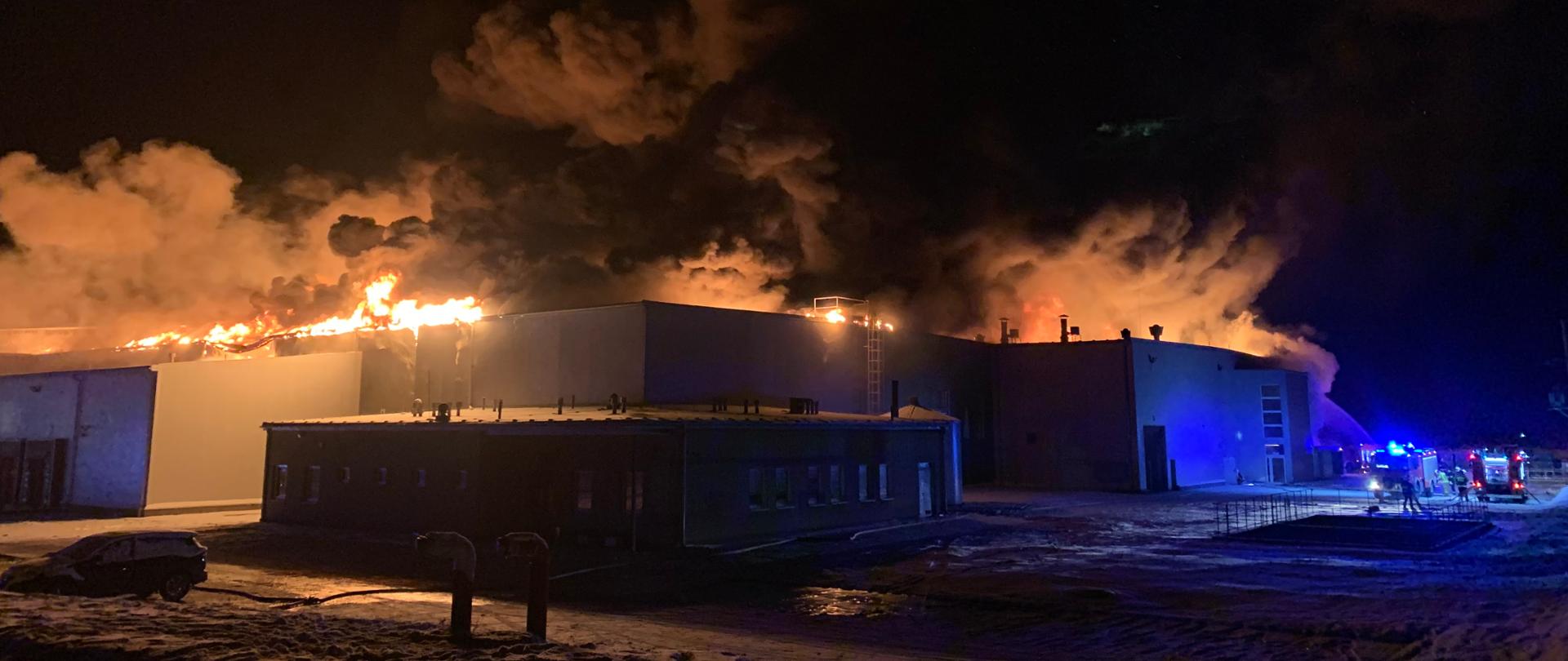 Zdjęcie zrobione w nocy, które przedstawia palącą się halę produkcyjną (perspektywa z zewnątrz). U góry hali widać wydostające się języki ognia oraz ogromne kłęby dymu. Z prawej strony zdjęcia widać w oddali trzy samochody strażackie oraz strażaków podczas akcji.