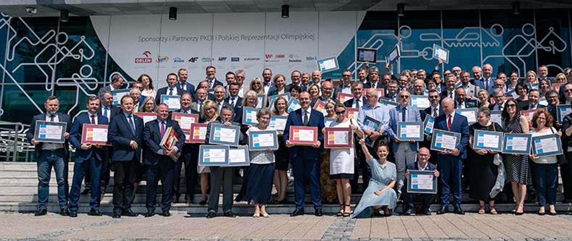 Grupa kilkudziesięciu osób, które trzymają w rękach duże dyplomy w ramkach. 