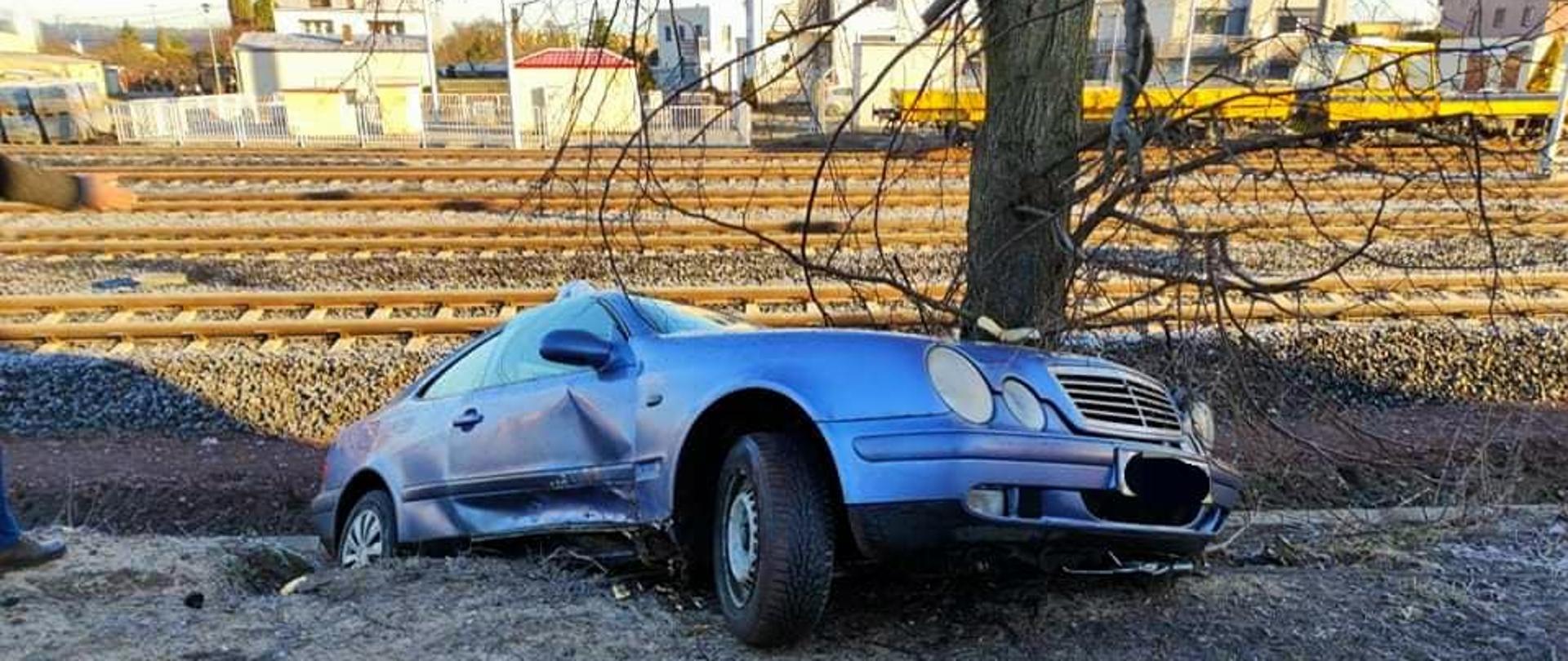 Widać uszkodzony samochód, który lewym bokiem znajduje się przy drzewie