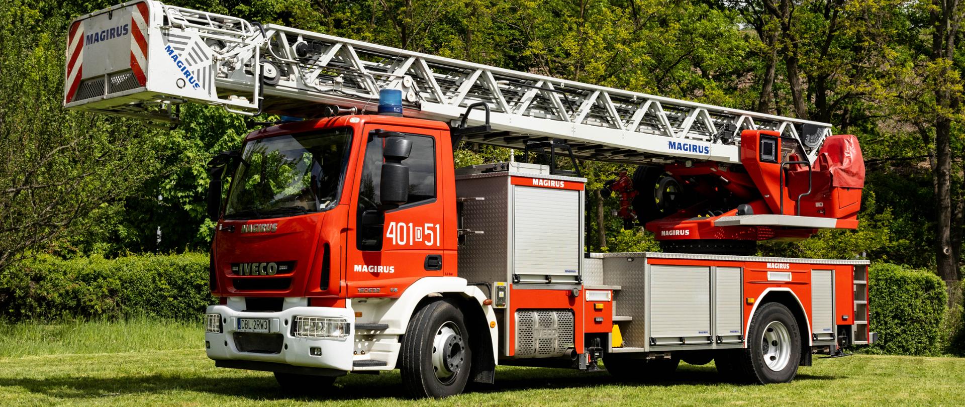 Samochód specjalny z drabiną mechaniczną marki IVECO Magirus oznaczony numerem „401[D]51”. Na podwoziu zamontowana jest długa, złożona drabina ratownicza z koszem na końcu, sygnowana logo „MAGIRUS”. Pojazd stoi na trawiastym tle.