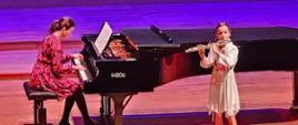 Dziewczynka gra na flecie na estradzie sali koncertowej, za nią kobieta gra na fortepianie.