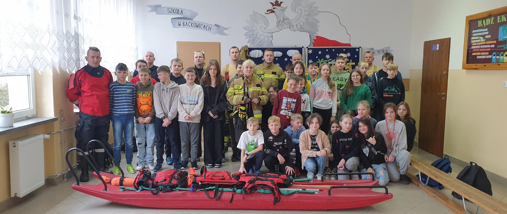 Zdjęcie grupowe strażaków z państwowej i ochotniczej straży pożarnej wraz z uczniami na korytarzu szkoły, przed wszystkimi na podłodze sanie lodowe wraz ze sprzętem do ratownictwa wodnego