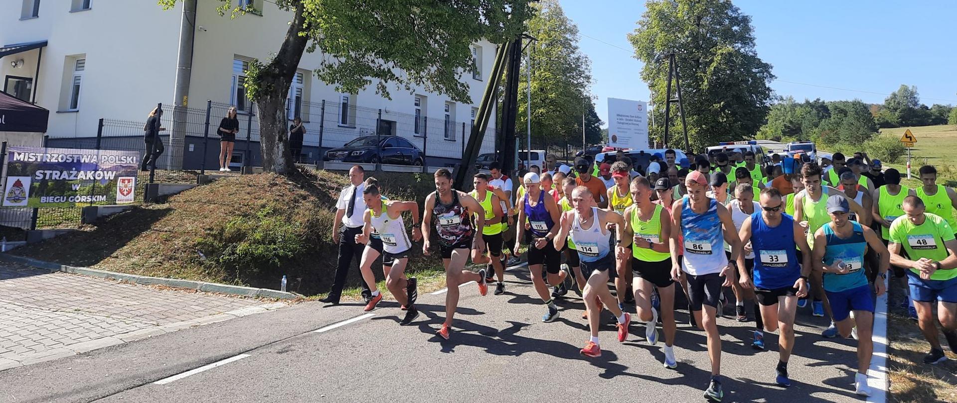 Na zdjęciu znajdują się strażacy startujący do biegu. W tle drzewa i budynek 