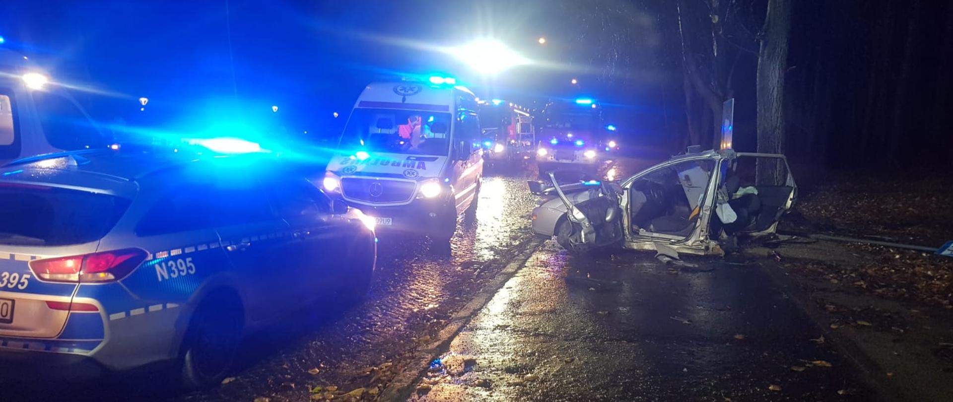 Zdjęcie ze zniszczonym przodem przełamanego samochodu w tle samochód straży pożarnej z włączonymi niebieskimi sygnałami i strażacy, samochody policji oraz karetki pogotowia.
