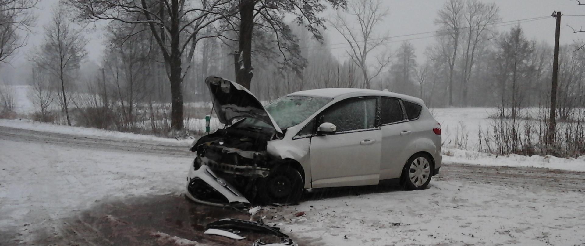 Uszkodzony srebrny samochód osobowy marki Ford stojący na zaśnieżonej drodze. Pojazd ma uszkodzony przód - zerwany zderzak, zniszczony pas przedni, podniesiona pogięta klapa przedniej maski. Przed pojazdem połamane elementy karoserii. Widok pojazdu od strony pasażera.