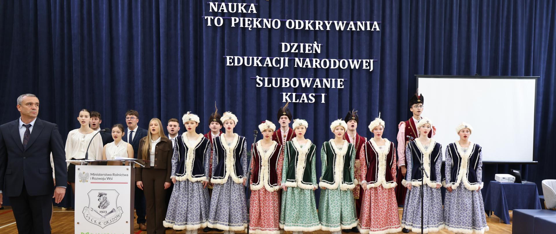 Uczniowie podczas szkolnej uroczystości Dnia Edukacji Narodowej i ślubowania klas pierwszych występują na scenie w strojach ludowych.