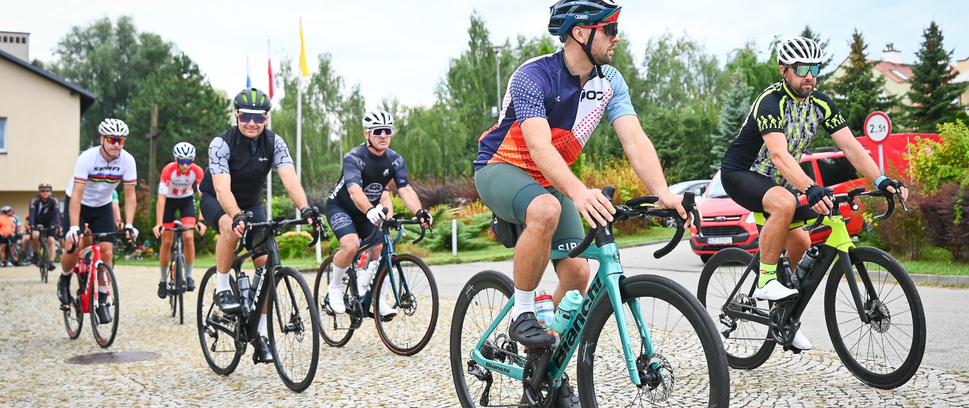 Grupa kolarzy-strażaków rusza na trasę pielgrzymki, jadąc po kostce brukowej przed kościołem. Na pierwszym planie rowerzysta w sportowym stroju z elementami niebieskiego i pomarańczowego oraz białym kasku. Za nim jadą kolejni uczestnicy. W tle widoczny jest budynek kościoła, flagi i czerwony samochód strażacki.