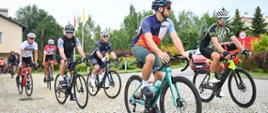 Grupa kolarzy-strażaków rusza na trasę pielgrzymki, jadąc po kostce brukowej przed kościołem. Na pierwszym planie rowerzysta w sportowym stroju z elementami niebieskiego i pomarańczowego oraz białym kasku. Za nim jadą kolejni uczestnicy. W tle widoczny jest budynek kościoła, flagi i czerwony samochód strażacki.