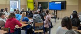 Pracownik Wojewódzkiego Inspektoratu Ochrony Środowiska w Warszawie prezentuje slajd, kilkudziesięcioro uczniów siedzi w ławkach w szklonej klasie.