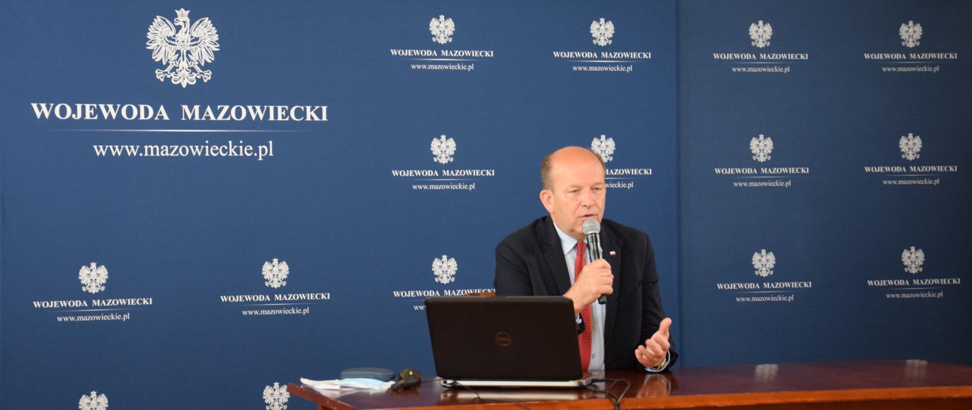 Na zdjęciu znajduje się mężczyzna siedzący przed laptopem mówiący do mikrofonu