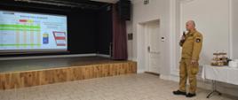 zdjęcie przedstawia strażaka w ubraniu służbowym z mikrofonem podczas referowania, w tle widoczny projektor