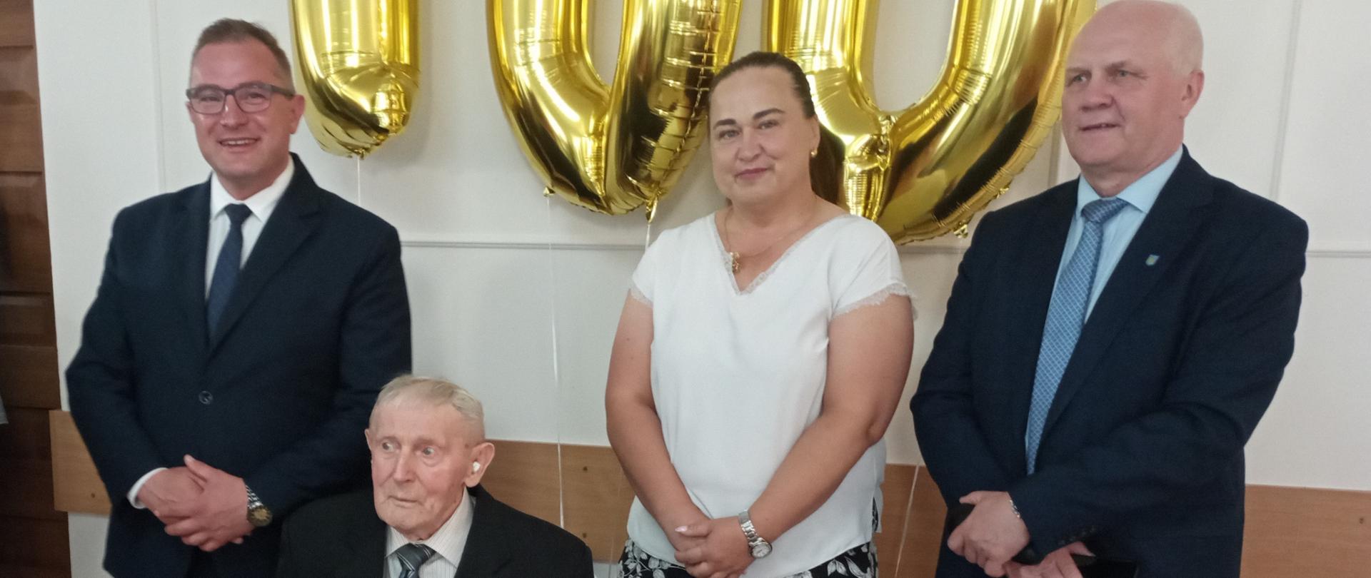 Starszy mężczyzna siedzi na krześle trzymając w rękach zdobioną drewnianą laskę. Po jego lewej stronie stoi mężczyzna a po prawe -kobieta i mężczyzna. Z tyłu nad nimi wiszą złote balony w kształcie liczby 100. Obok stoi ogromny kosz kwiatów i paczki z prezentami.