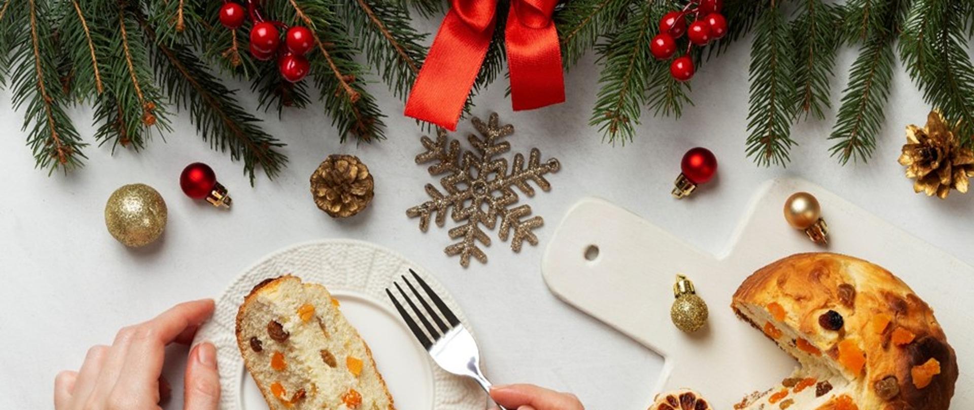 U góry zielone gałązki jedliny z kolorowymi bombkami. Po prawej stronie na białej desce leży ciasto z bakaliami. Po lewej dłoń trzymająca widelec oraz biały talerz z kawałkiem ciasta na białym talerzu.