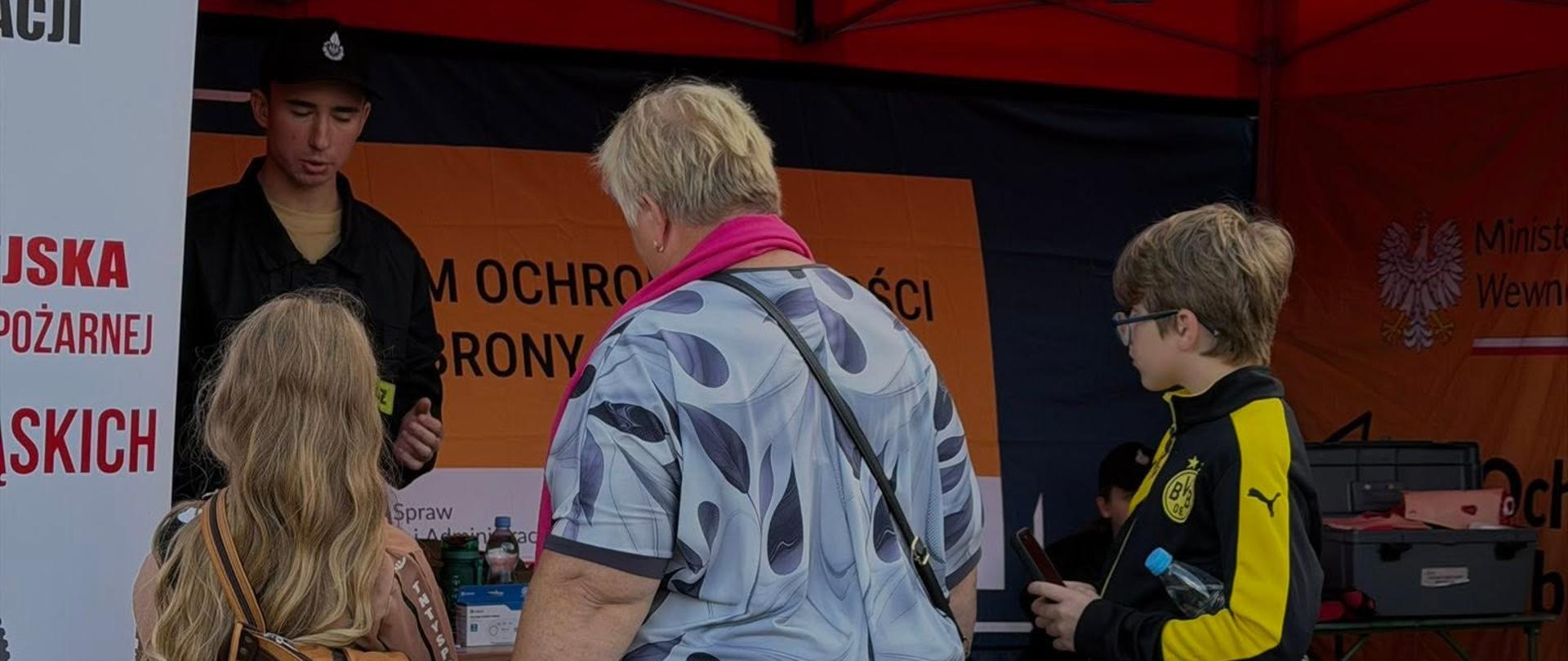 Na zdjęciu przedstawiono stoisko edukacyjne poświęcone ochronie ludności i obronie cywilnej. Odwiedzający rozmawiają z przedstawicielem straży pożarnej oraz zapoznają się z materiałami dotyczącymi bezpieczeństwa i przygotowania na sytuacje kryzysowe. W namiocie widoczne są materiały informacyjne Programu Ochrony Ludności i Obrony Cywilnej oraz fantomy wykorzystywane do nauki udzielania pierwszej pomocy.