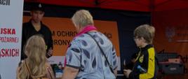 Na zdjęciu przedstawiono stoisko edukacyjne poświęcone ochronie ludności i obronie cywilnej. Odwiedzający rozmawiają z przedstawicielem straży pożarnej oraz zapoznają się z materiałami dotyczącymi bezpieczeństwa i przygotowania na sytuacje kryzysowe. W namiocie widoczne są materiały informacyjne Programu Ochrony Ludności i Obrony Cywilnej oraz fantomy wykorzystywane do nauki udzielania pierwszej pomocy.