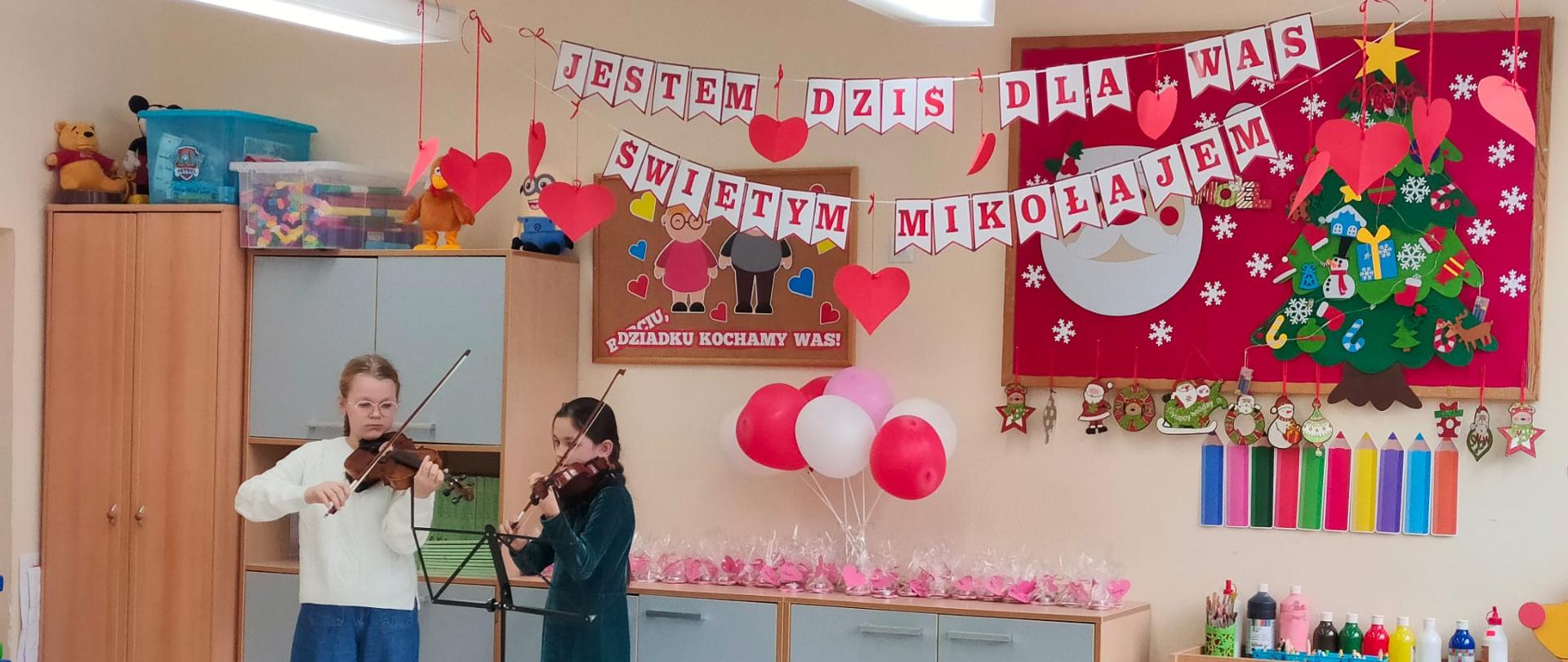 Po lewej stronie na zdjęciu Alicja Kosytorz i Laura Luchowska (duet skrzypcowy) podczas występu z okazji Dnia Babci i Dziadka w ZS-P nr 5 w Żaganiu. Po prawej stronie kolorowy napis: Jestem dziś dla Was Świętym Mikołajem. Dekoracja z serduszek i kolorowych balonów.