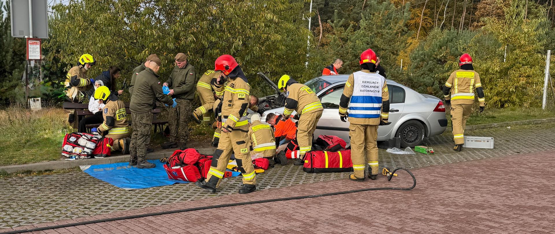 Zdjęcie przedstawia scenę pozorowanego wypadku lub ćwiczeń ratownictwa drogowego, gdzie grupa strażaków (część z nich w hełmach i ubraniach bojowych) oraz inni ratownicy zajmują się srebrnym samochodem osobowym na parkingu. Widać, jak ratownicy udzielają pomocy w okolicach pojazdu, leżą na ziemi plandeki i sprzęt medyczny/ratunkowy, a jedna kobieta w cywilnym ubraniu obserwuje akcję. W tle widoczna jest roślinność, drzewa oraz słup energetyczny.