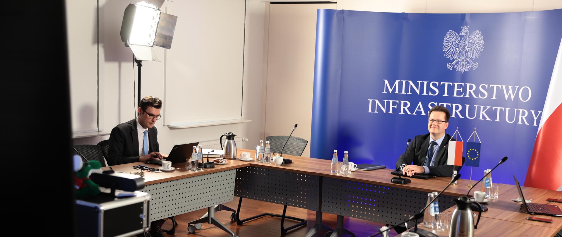 Na zdjęciu widać dwóch mężczyzn siedzących przy stole konferencyjnym. W tle jednego z mężczyzn stoi ścianka z napisem Ministerstwo Infrastruktury. Po bokach stoją lampy oświetlające pomieszczenie.
