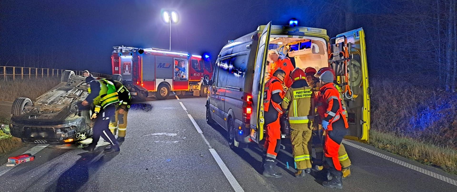 Zdjęcie przedstawia teren działań po zderzeniu dwóch samochodów osobowych na drodze krajowej nr 65. Z lewej strony znajduje się pojazd osobowy usytuowany na dachu (przy nim znajdują się strażak i policjant). Na środku obrazu widać pojazd pożarniczy oświetlający miejsce akcji. Po prawej stronie można zaobserwować ratowników medycznych i strażaków, którzy lokują osobę poszkodowaną do ambulansu.