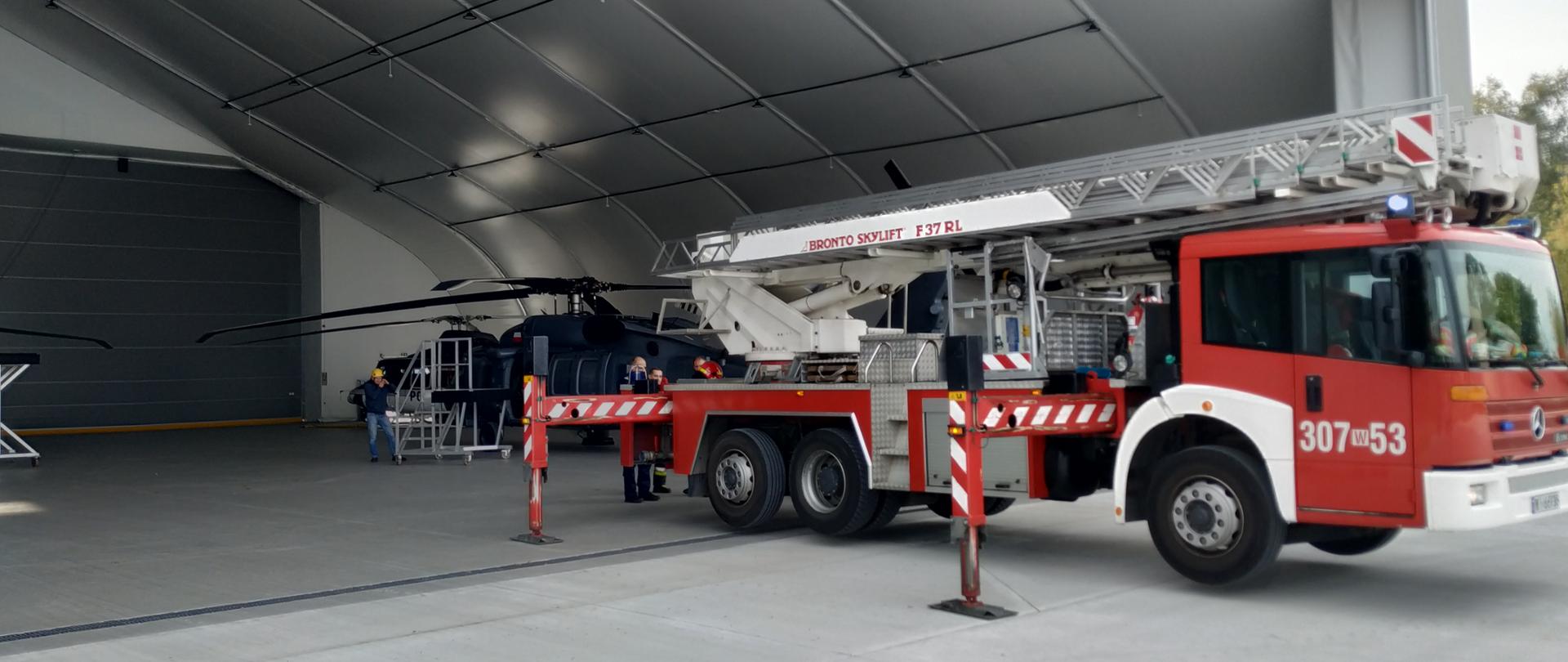 Na pierwszym planie zdjęcia widać strażacki podnośnik hydrauliczny. W tle zdjęcia widać częściowo wnętrze hangaru typu lekkiego oraz szary śmigłowiec.