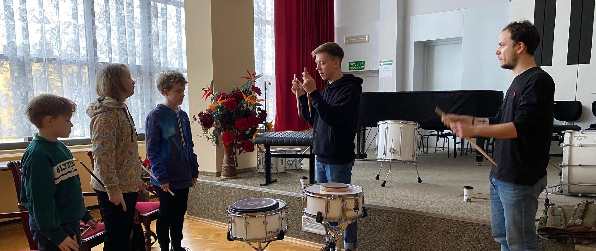 Kacper Kałuża i Adam Barański z trójką uczniów trzymających w rękach pałki na auli PSM, w tle scena