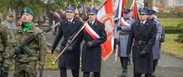 Poczty sztandarowe maszerują w uroczystym przemarszu. Funkcjonariusze w granatowych i szarych mundurach niosą biało-czerwone sztandary z godłem Polski. Obok nich idzie żołnierz w zielonym beretcie. W tle widoczne są kolejne poczty oraz uczestnicy uroczystości w jesiennym parku.