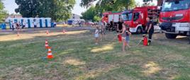 Zdjęcie przedstawia strażaków podczas festynu. Dzieci przy pomocy hydronetek strącają butelki z pachołków. Na zdjęciu widać również samochody strażackie