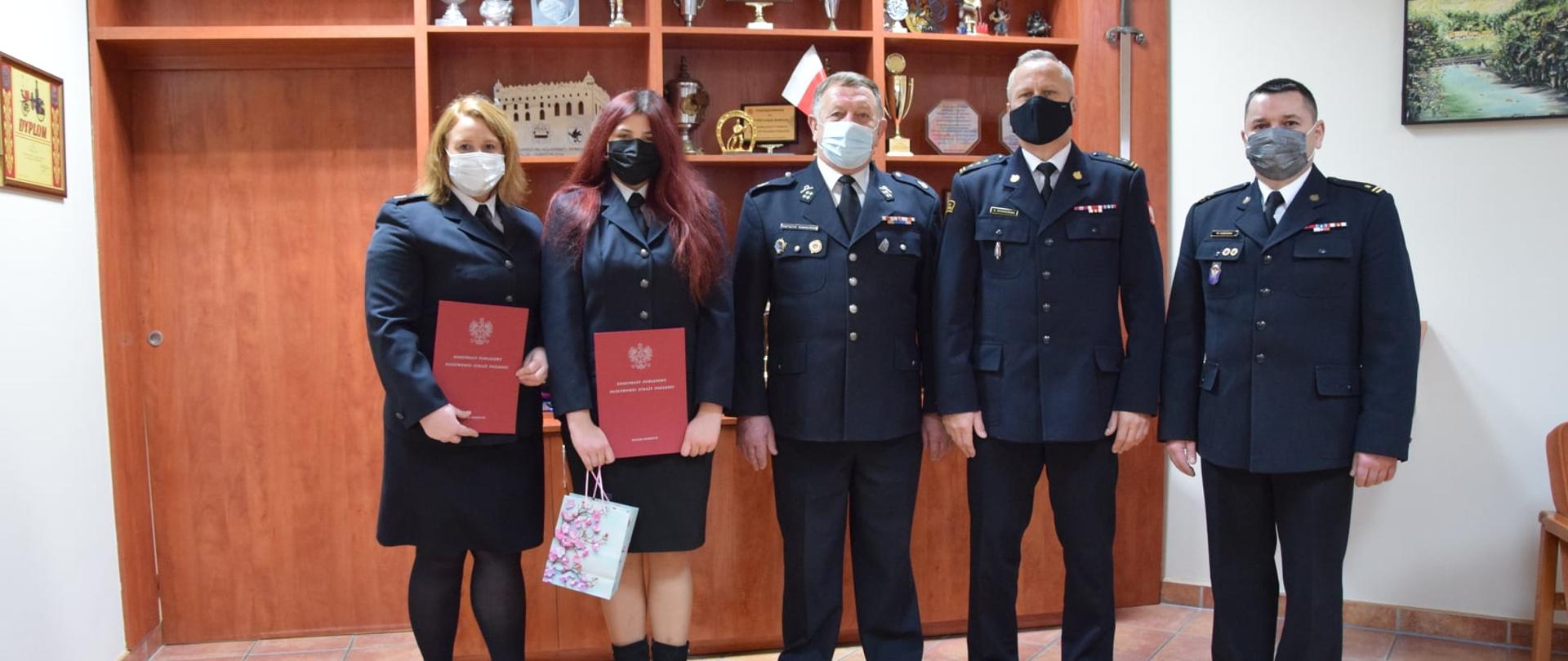 Od lewej strony stoją druhny OSP Wrocki, jedna z druhem trzyma w dłoniach paczuszkę i list gratulacyjny w bordowej teczce, dalej stoi druh OSP Wrocki a za nim Zastępca Kujawsko-Pomorskiego Komendanta Wojewódzkiego PSP st. bryg. Robert Wiśniewski wraz z Komendantem Powiatowym PSP w Golubiu-Dobrzyniu. Wszyscy strażacy na zdjęciu są w umundurowaniu galowym.