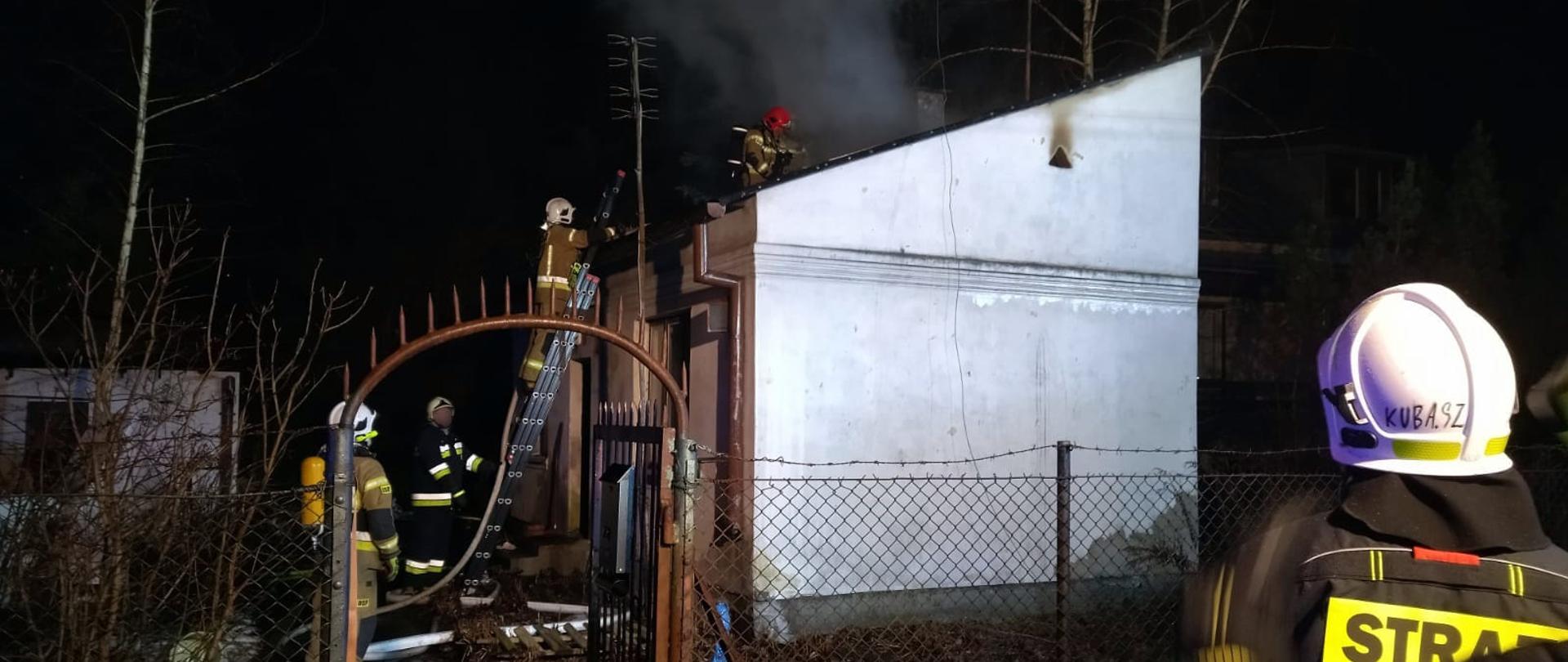 Strażacy prowadzą działania gaśnicze na dachu budynku mieszkalnego. Jeden strażak znajduje się na dachu drugi stoi na drabinie i podtrzymuje linie gaśniczą.