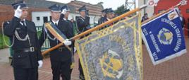 Jubileusz 95-lecia OSP Konary. Plac przed remizą strażacką. Poczty sztandarowe oddające honor w trakcie hymnu państwowego. W tle budynki. 