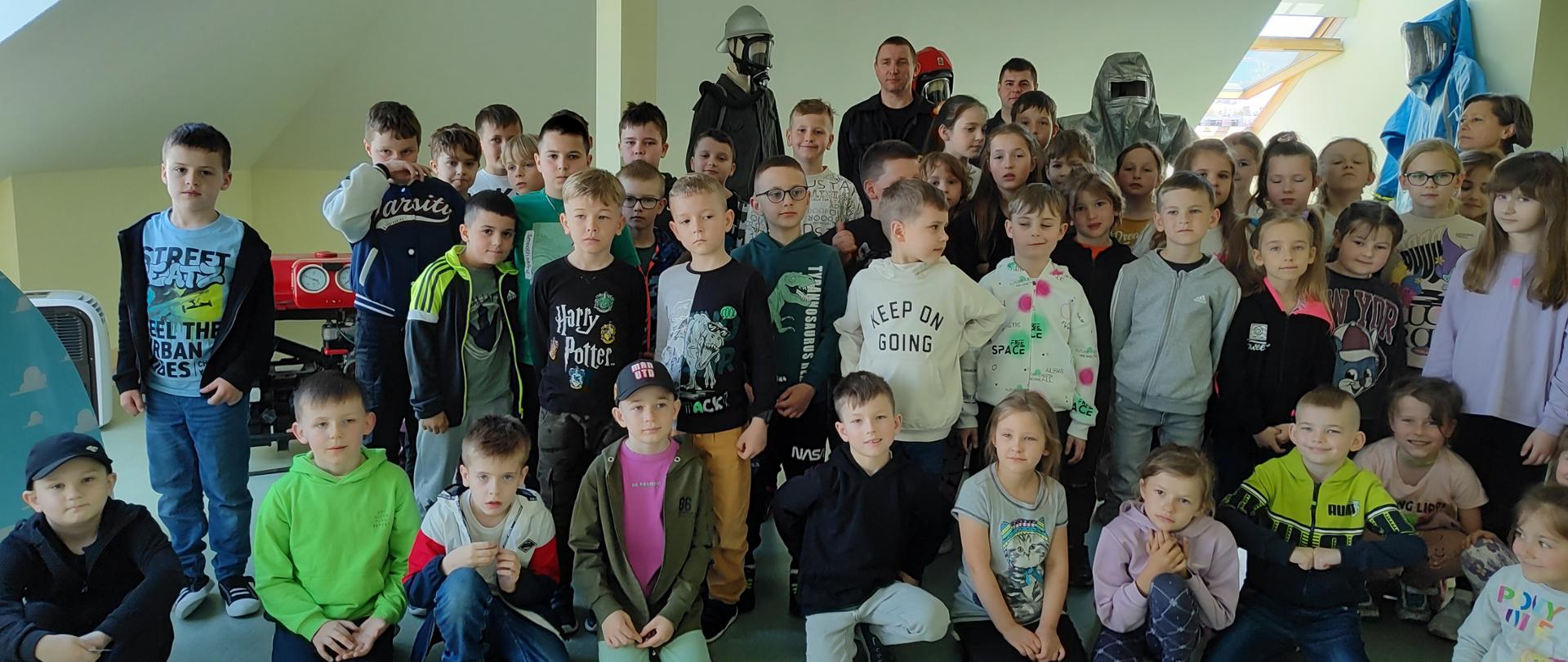 Zdjęcie grupowe na tle ubrań bojowych strażackich: przeciwgazowego, żaroochronnego oraz ubrania specjalnego starego i nowego typu. Po lewej stronie widać motopompę pożarniczą. 