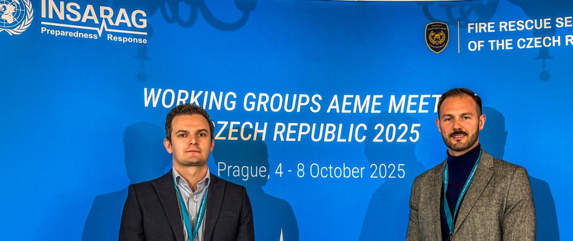 Dwóch mężczyzn pozuje na tle ścianki konferencyjnej z logiem INSARAG i napisem "Working Groups AEME Meeting, Czech Republic 2025". Obaj mają identyfikatory na smyczach i są ubrani w styl formalno-biznesowy. Zdjęcie wykonane w miejscu spotkania w Pradze.