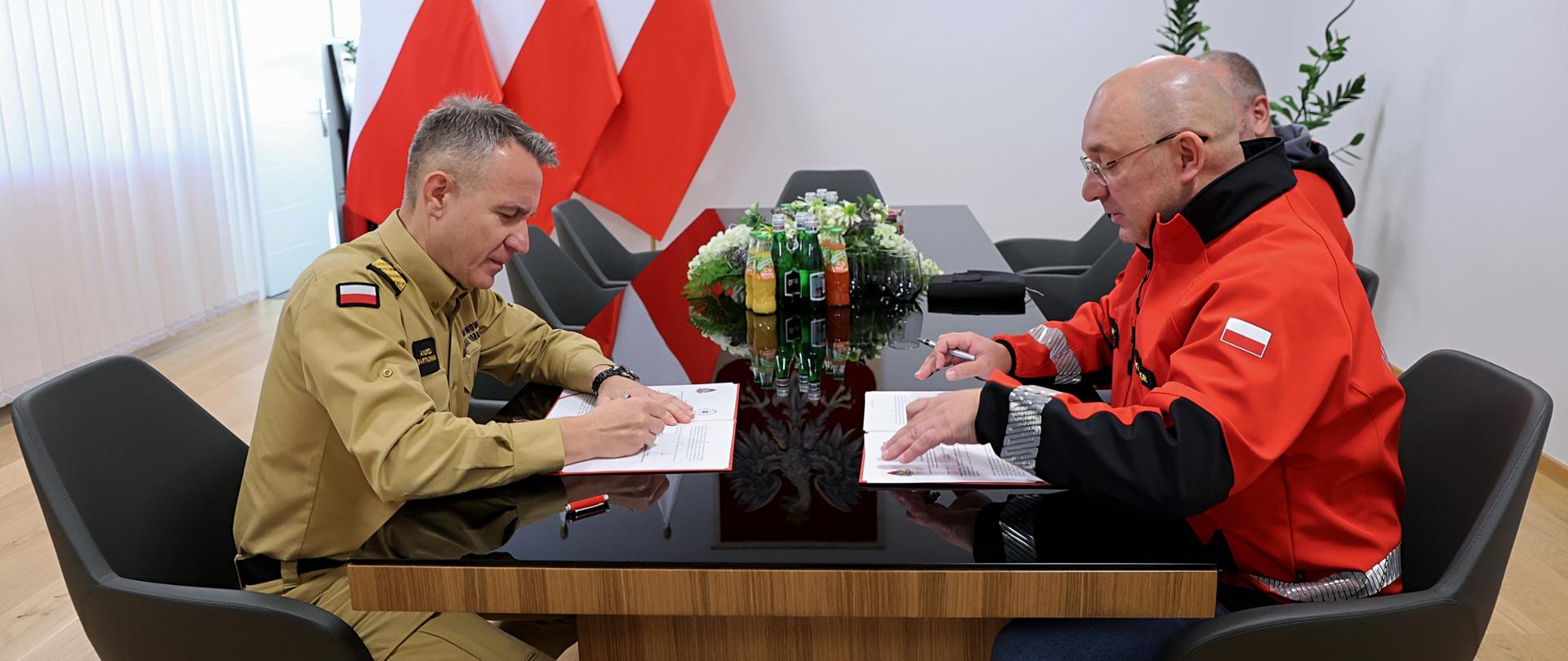 Komendant główny PSP w umundurowaniu służbowym w kolorze piaskowym i dyrektor LPR w ubraniu w kolorze czerwonym z napisem: Lotnicze Pogotowie Ratunkowe siedzący przy stole w trakcie podpisywania porozumienia. 