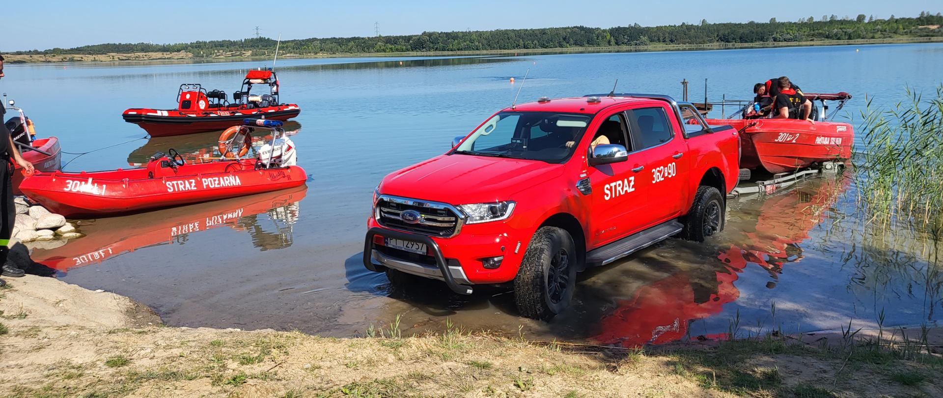 Samochód strażacki w trakcie wodowania łodzi ratowniczej, na drugim planie dwie zwodowane łodzie strażackie