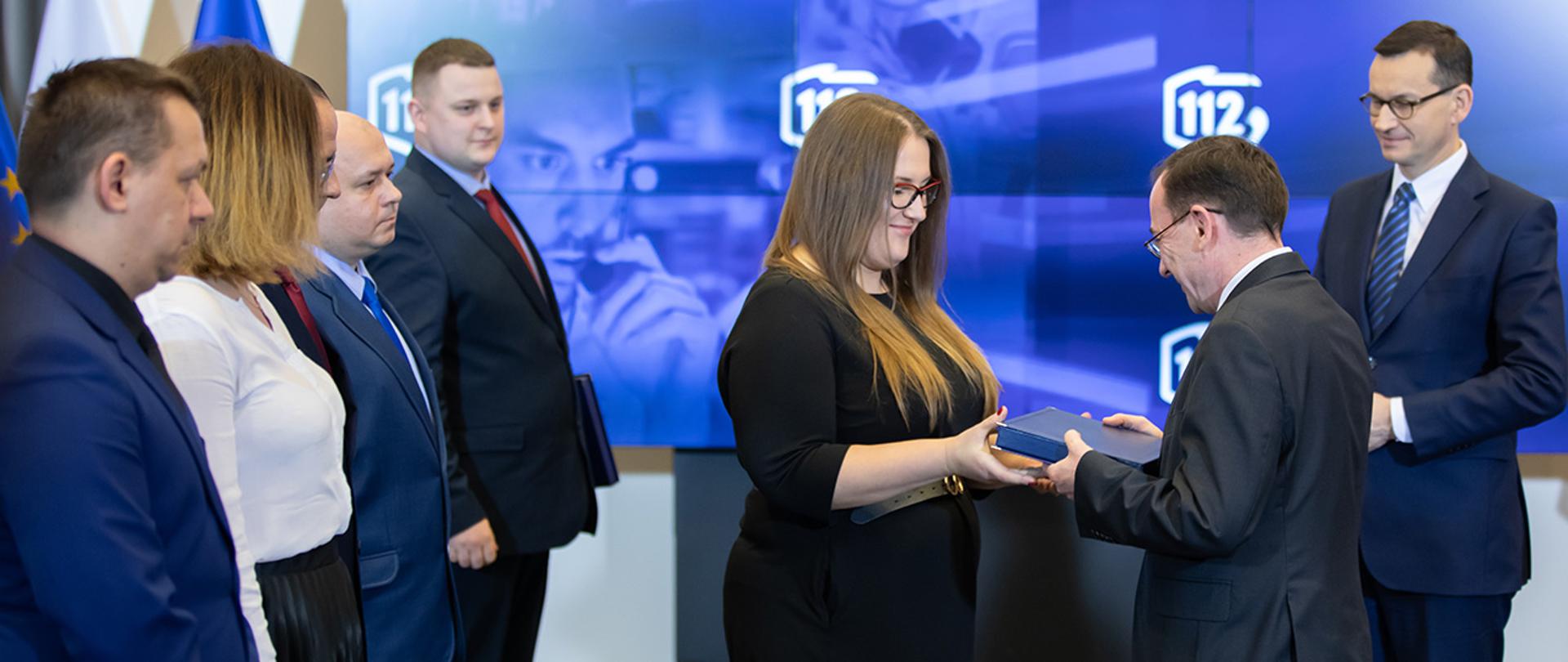 Na zdjęciu widać jak minister Mariusz Kamiński wręcza podziękowanie jednej z pracownic CPRu. Widać również premiera Mateusza Morawieckiego i innych pracowników CPR.