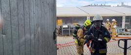 Na pierwszym planie widać strażaków w sprzęcie ochrony układu oddechowego wchodzących z linią wężową do budynku warsztatowego.